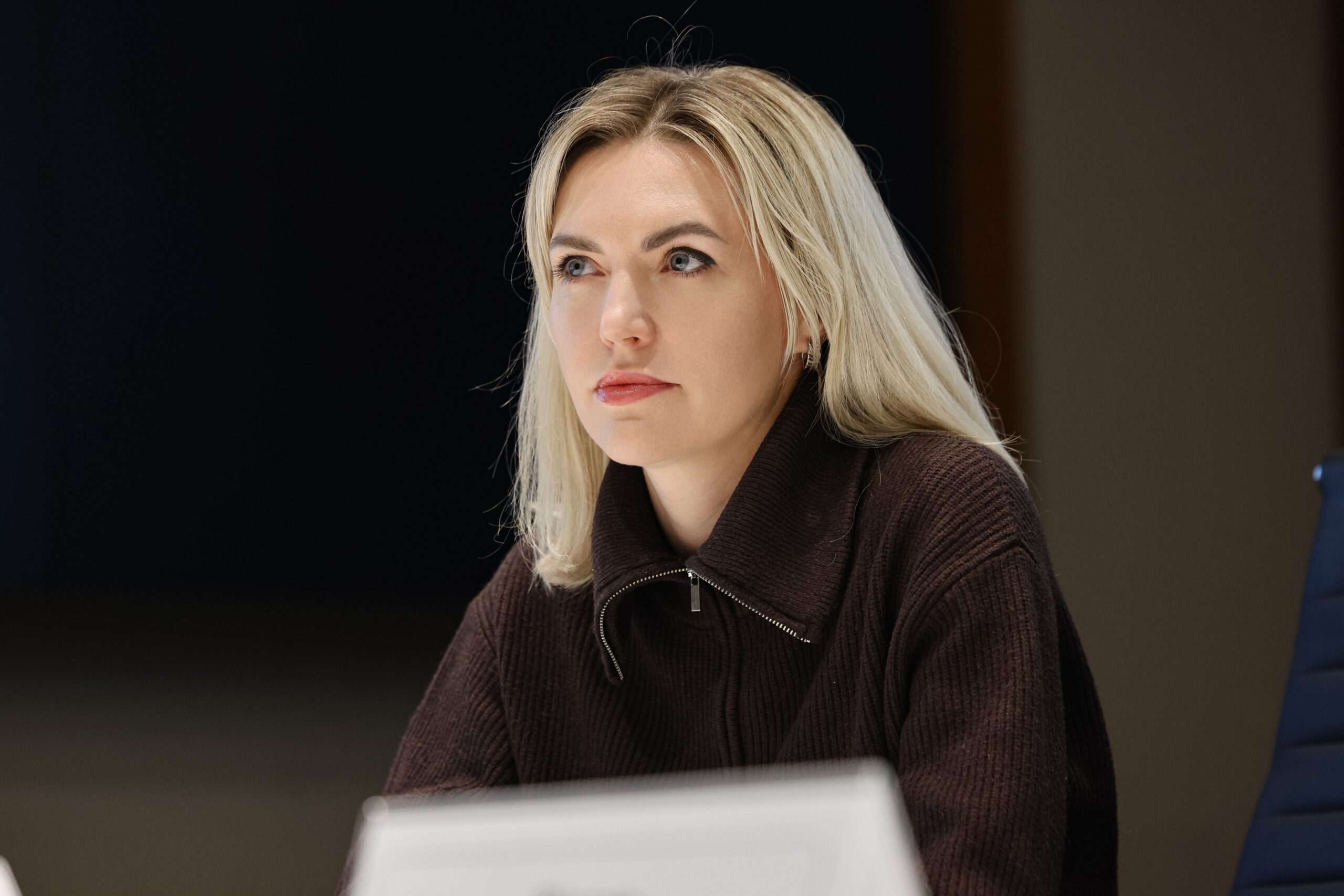 Жінка зі світлим волоссям у коричневому светрі на блискавці сидить за столом, зосереджено дивлячись убік під час офіційного заходу. Перед нею на столі розміщена ідентифікаційна табличка, а фон залишається темним і розмитим.