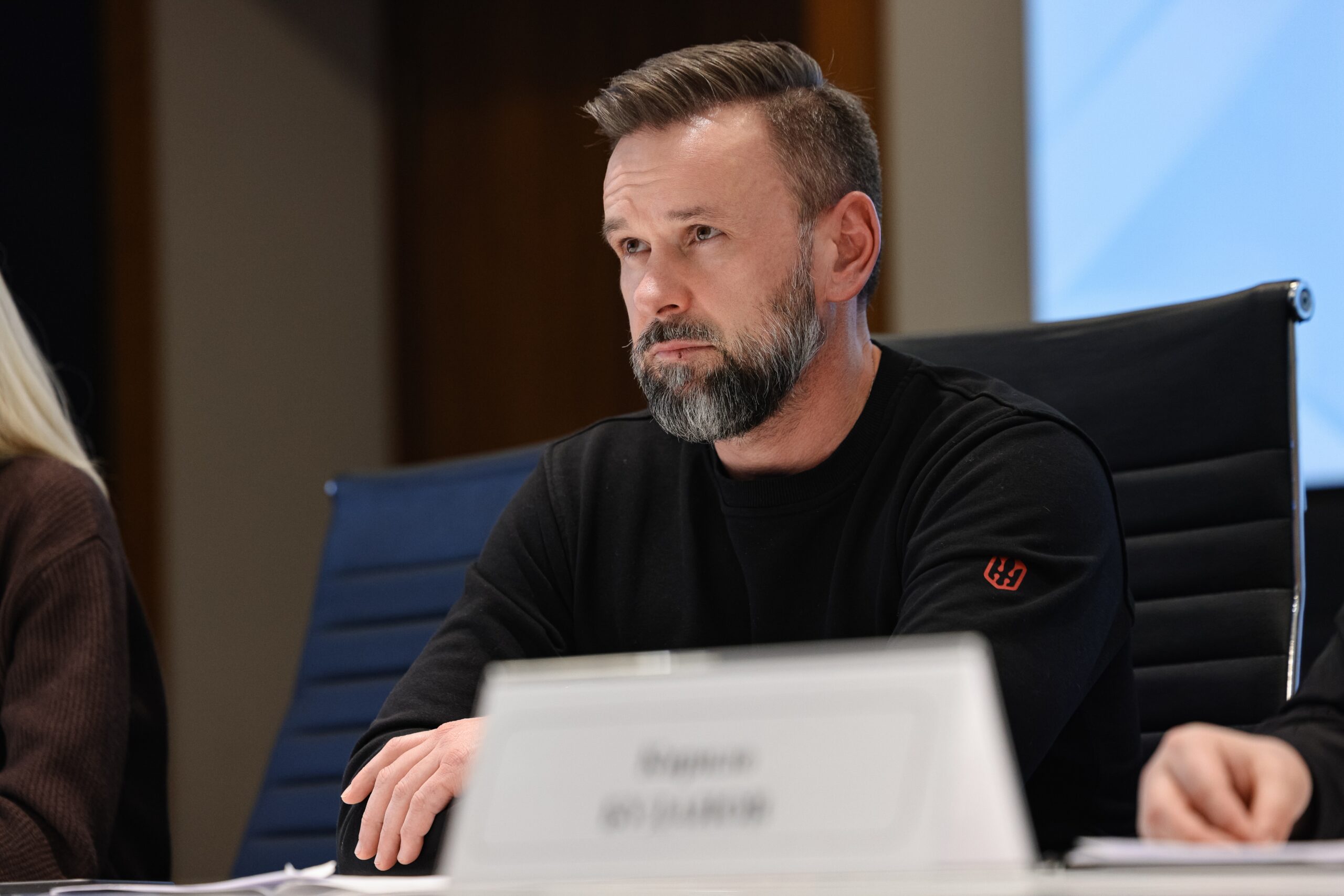 Чоловік із сивіючою бородою та в чорному світшоті сидить за столом у конференц-залі, зосереджено дивлячись убік. Перед ним на столі стоїть іменна табличка, а на задньому плані видніється частина синього екрана офіційного заходу.