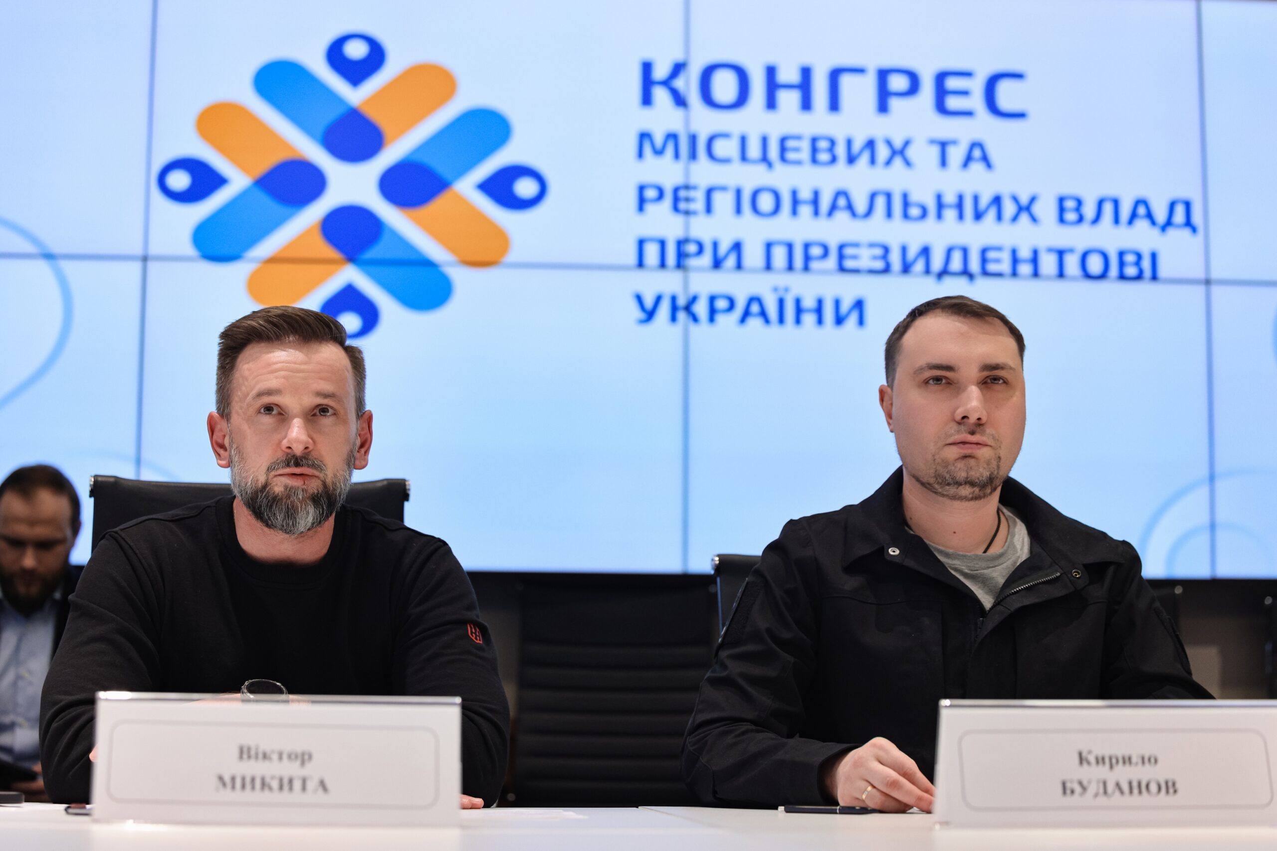 Двоє чоловіків у темному одязі сидять за столом під час засідання на фоні великого синьо-білого екрана з логотипом та офіційним написом. Перед кожним із них на столі розміщена ідентифікаційна табличка з прізвищем, а їхні погляди зосереджені на подіях заходу.