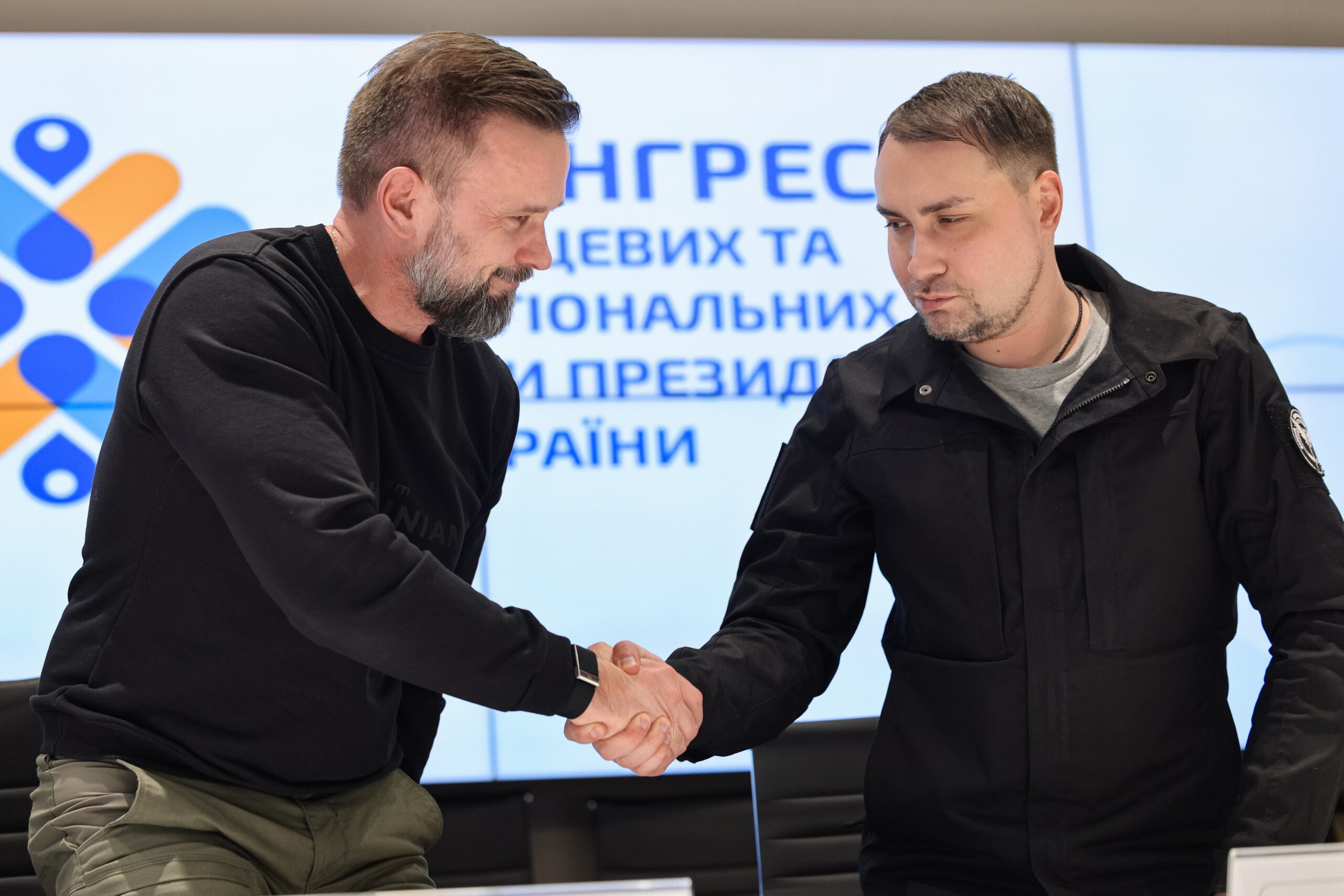 Двоє чоловіків у темному одязі стоять перед офіційним екраном і потискають один одному руки. Вони дивляться один на одного зі стриманими усмішками, а на задньому плані видніється напис про засідання Конгресу місцевих та регіональних влад.