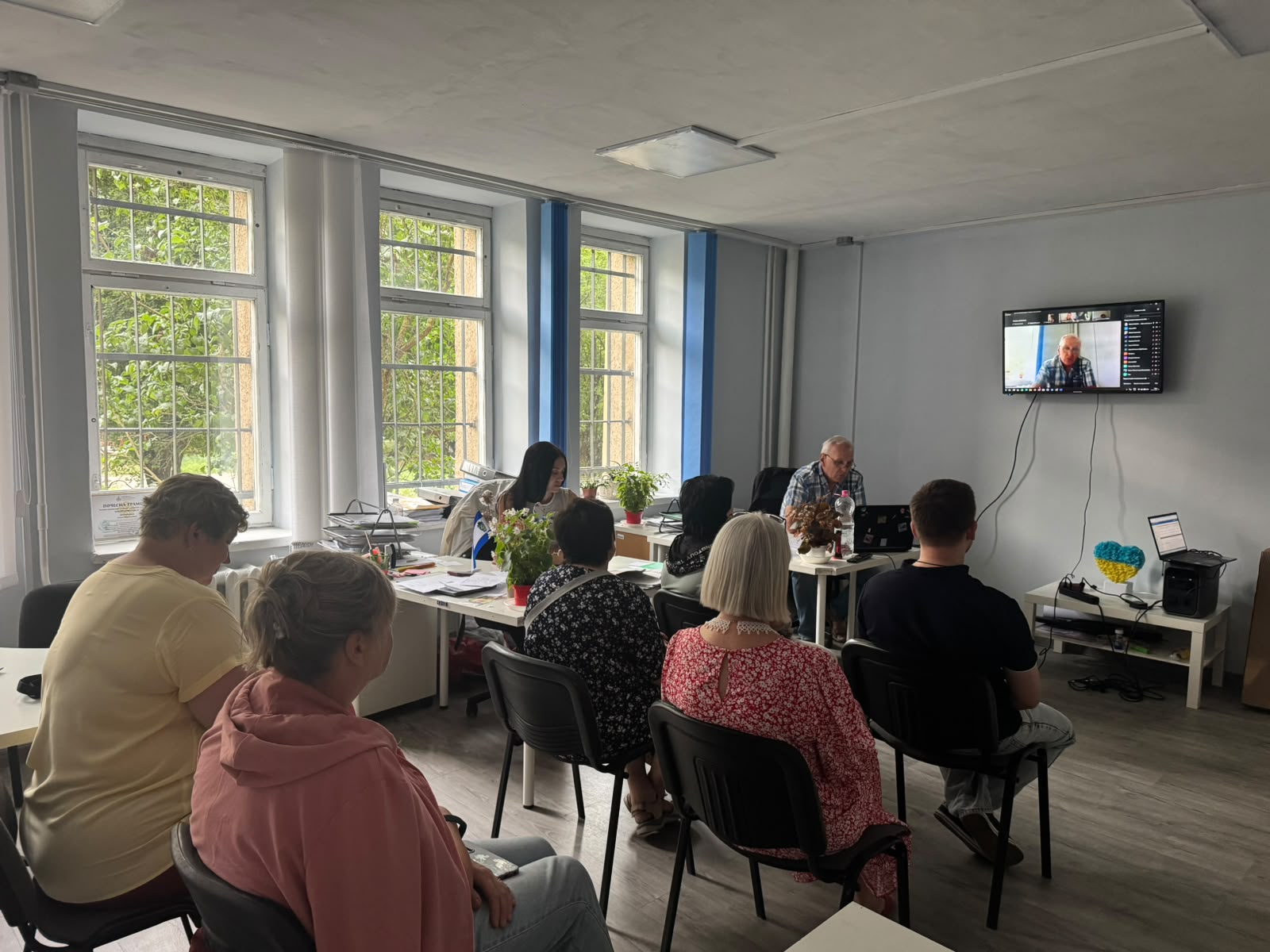 Група людей у світлому кабінеті гуманітарного хабу бере участь в онлайн-нараді або вебінарі. Присутні сидять на стільцях спиною до камери та уважно дивляться на настінний телевізор, де транслюється виступ спікера через відеозв'язок. За робочими столами біля вікна сидять співробітники хабу з ноутбуком та робочими документами. Фотографія 3