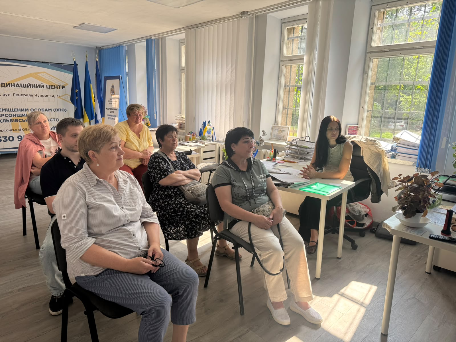 Учасники зустрічі та співробітниця хабу сидять у приміщенні координаційного центру та слухають виступ спікера. У лівому кутку кімнати встановлено державний прапор України, прапор ЄС та інформаційний банер Координаційного центру допомоги ВПО з Херсонщини. Біля вікна за столом із паперами сидить темноволоса фахівчиня.