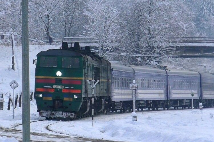 Пасажирський потяг із зеленим локомотивом рухається засніженою залізничною колією серед засніжених дерев і пагорбів у зимовий день.