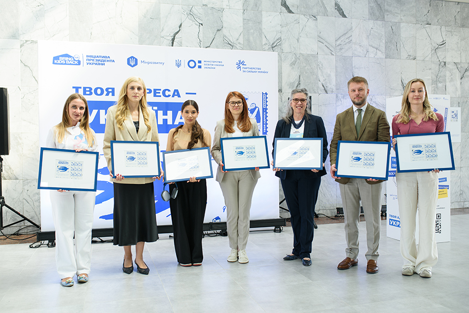 Семеро учасників офіційної презентації (п'ять жінок і двоє чоловіків у діловому одязі) вишикувалися в ряд у холі з мармуровими стінами перед тематичним банером. Кожен із присутніх тримає перед собою поштовий блок марок спецпогашення «Твоя адреса — Україна» в темно-синій дерев'яній рамці.