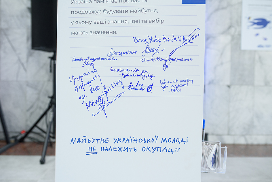 Білий інформаційний стенд із друкованим закликом «МАЙБУТНЄ УКРАЇНСЬКОЇ МОЛОДІ НЕ НАЛЕЖИТЬ ОКУПАЦІЇ» та рукописними словами підтримки й підписами, залишеними синім маркером. Серед авторів послань — представники ініціативи «Bring Kids Back UA», Мінрозвитку, Мінсоцполітики, а також Посольств Канади та Великої Британії.