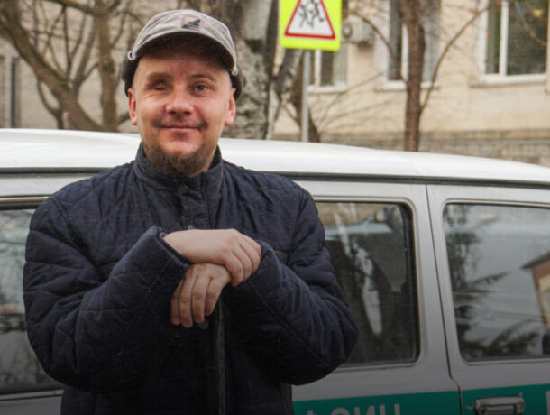 Чоловік у стьобаній темно-синій куртці та світлій кепці, усміхається, стоячи біля світлого автомобіля (ймовірно, службового) на вулиці міста. Позаду видно дорожній знак 'Діти' та фасади будівель.