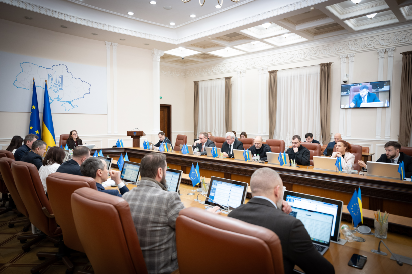 Загальний план робочої наради у великій світлій залі. Учасники сидять за П-подібним столом із ноутбуками та мікрофонами. На стіні розміщено контурну карту України з гербом та державні прапори, а на екрані телевізора триває відеозв'язок.