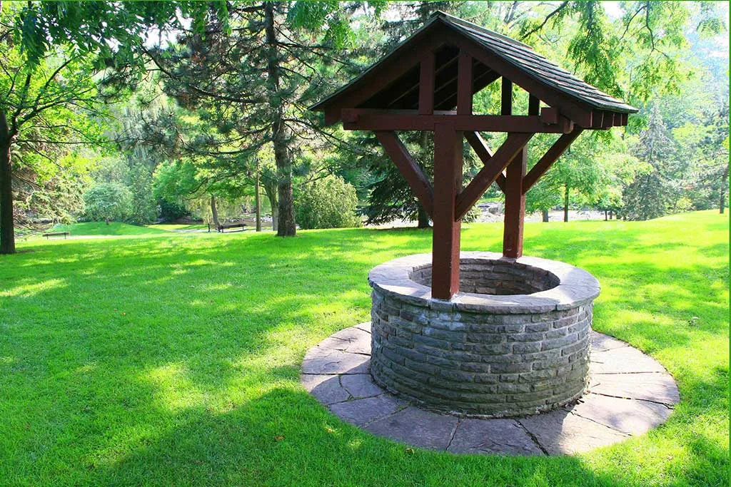 Кам'яний колодязь із дерев'яним дахом, розташований на доглянутому зеленому газоні. Навколо колодязя видно плити бруківки, а на задньому плані — густі дерева та сонячне світло.
