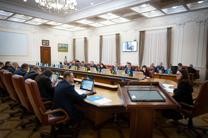 Група людей за круглим столом у великому залі засідань з класичним інтер'єром під час офіційної зустрічі.