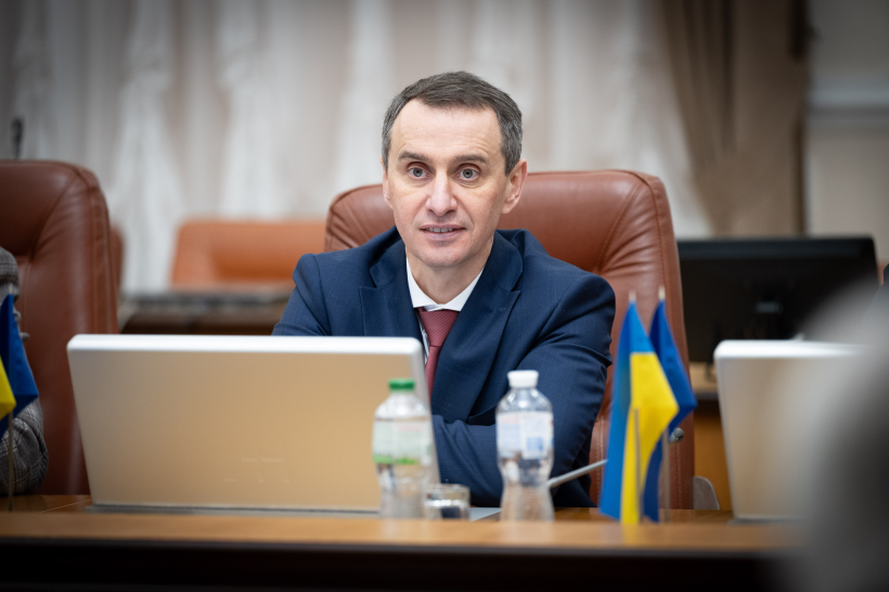 Портрет чоловіка у діловому костюмі та краватці, який виступає під час офіційної наради. Він сидить за робочим столом перед ноутбуком, на фоні світлого інтер'єру зали засідань.