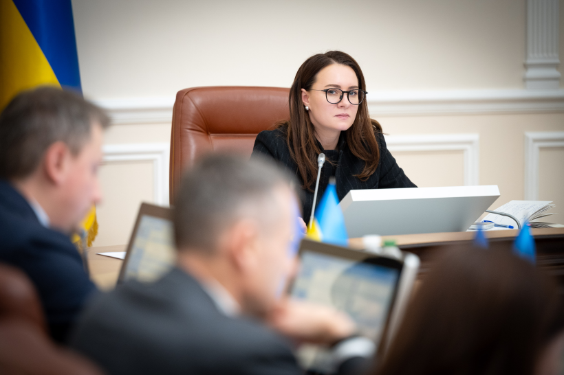 Жінка в окулярах та офіційному одязі сидить за столом під час наради, на фоні державного прапора України.