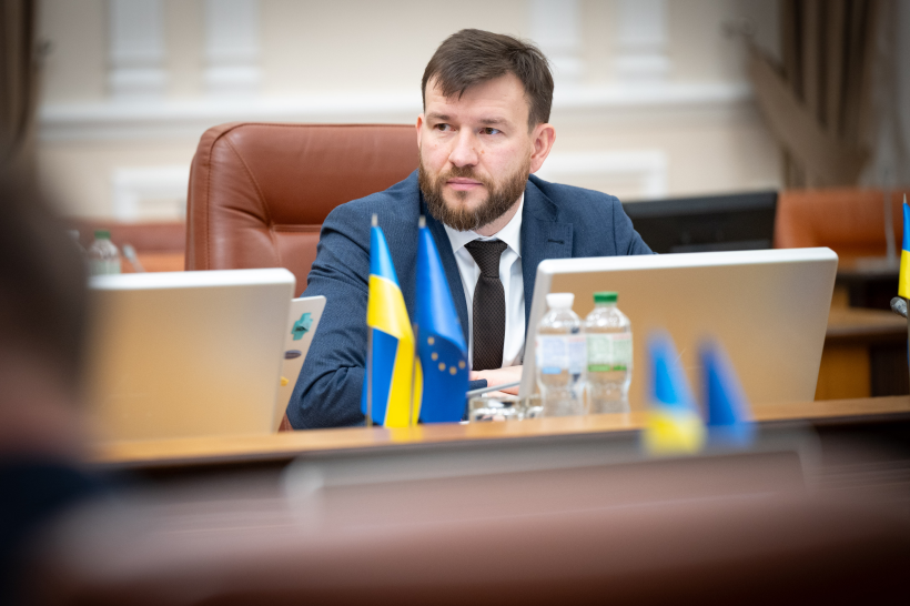 Чоловік у діловому костюмі сидить за столом під час засідання, на фоні маленьких прапорів України та Європейського Союзу.