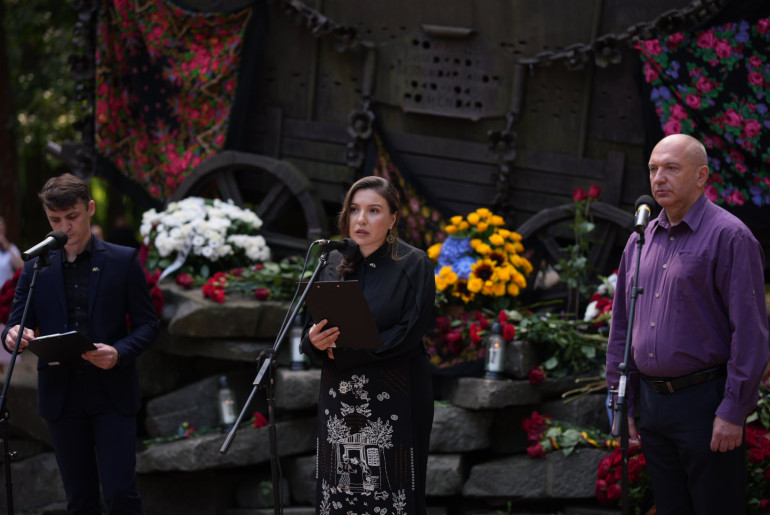 Троє людей виступають з промовами біля мікрофонів під час меморіального заходу на фоні пам’ятника у вигляді залізної кибитки. Монумент прикрашений квітами та традиційними хустками.