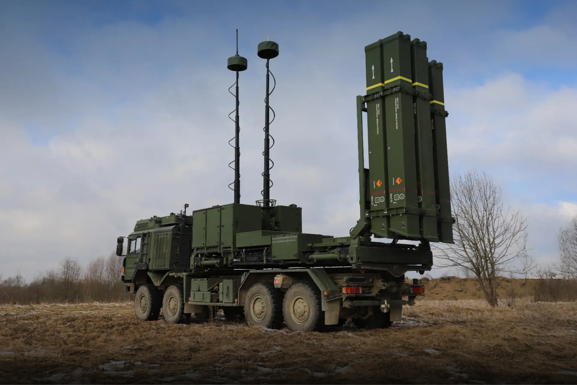 На відкритій місцевості під хмарним небом стоїть пускова установка зенітно-ракетного комплексу IRIS-T SLM на колісному шасі. Вертикальні контейнери для ракет підняті у бойове положення, поруч розгорнуті телескопічні щогли з антенами.