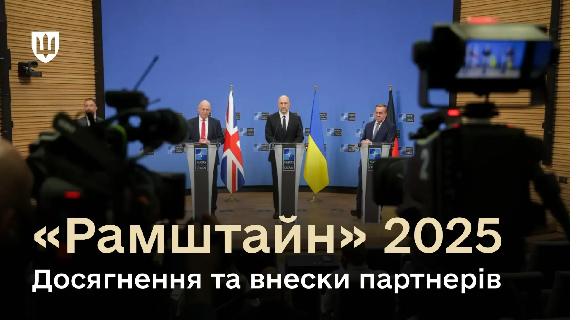 Троє офіційних представників стоять за трибунами на фоні прапорів Великої Британії, України та Німеччини під час пресконференції. На передньому плані видно силуети відеокамер.