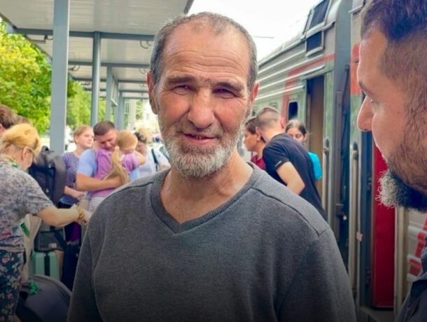 Літній чоловік з сивою бородою та зморшками, одягнений у сірий светр, посміхається, стоячи на пероні біля вагонів поїзда. На задньому плані видно людей, що сідають або виходять з поїзда.