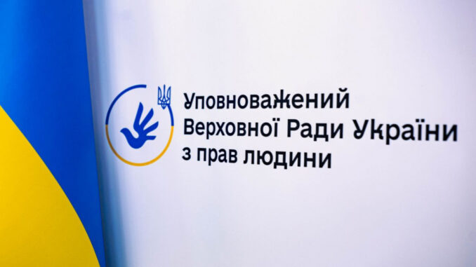 Офіційний логотип та назва інституції Омбудсмана України, розміщені на білому тлі. Зйомка виконана під невеликим кутом. У лівій частині кадру великим планом зафіксовано частину розгорнутого державного прапора України з насиченими синім та жовтим кольорами. Праворуч від прапора розташована офіційна символіка Уповноваженого: Кругла синьо-жовта емблема, всередині якої зображено синій силует розкритої долоні у вигляді птаха (голуба), що злітає. Зверху на контурі кола емблеми розміщено малий тризуб синього кольору. Праворуч від емблеми чітким чорним шрифтом у три рядки нанесено офіційну назву відомства: «Уповноважений Верховної Ради України з прав людини».
