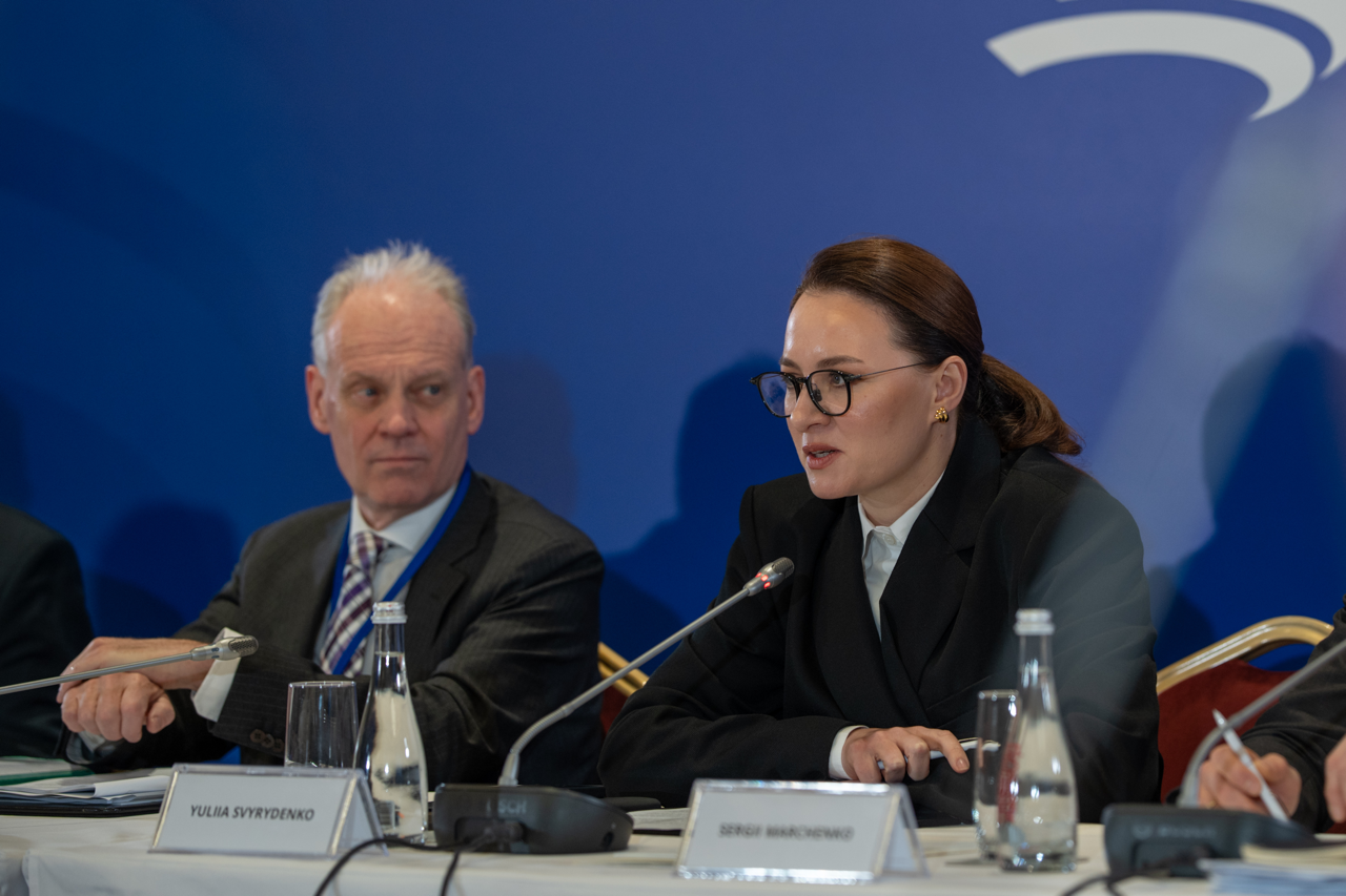 Жінка в окулярах та офіційному чорному костюмі виступає за столом з мікрофоном під час міжнародної конференції. Поруч із нею сидить чоловік у діловому костюмі, а перед ними встановлені таблички з іменами учасників та пляшки з водою на фоні синього банера.