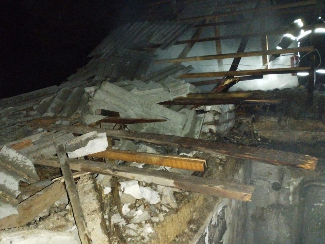 Зруйнований дах будівлі в нічний час. Видно пошкоджені дерев'яні балки, уламки шиферу та цегли. На задньому плані крізь конструкції пробивається дим і світло ліхтаря рятувальника.