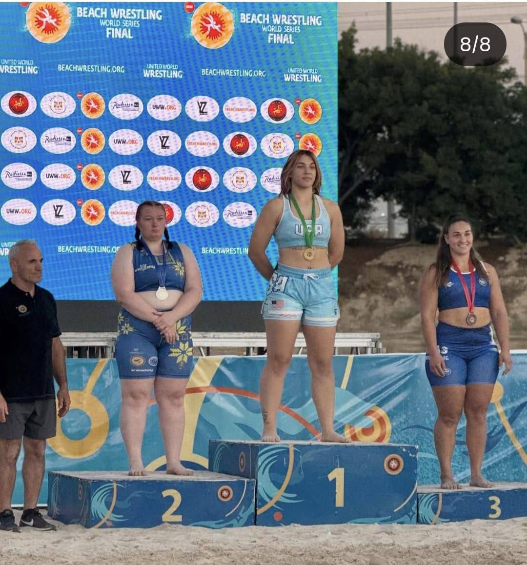 Церемонія нагородження на змаганнях з пляжної боротьби (Beach Wrestling World Series Final). Три спортсменки з медалями стоять на п'єдесталі переможців (№1, №2, №3) на піску. Переможниця (у центрі, №1, у блакитній формі з написом USA) стоїть на вищій сходинці. Зліва стоїть спортсменка, яка посіла друге місце (№2, у синій формі з жовтими елементами), а праворуч — третє місце (№3, у синій формі). Зліва на п'єдесталі стоїть чоловік у чорній футболці. На задньому плані видно великий екран з логотипами змагань та партнерів.
