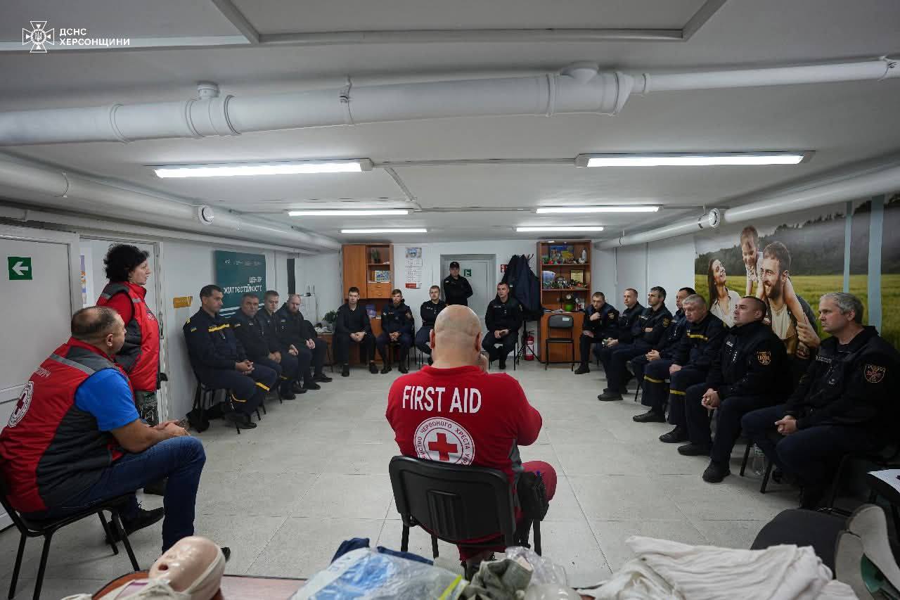Група з приблизно 20 людей, більшість з яких у формі ДСНС Херсонщини, сидить півколом у приміщенні (схожому на укриття або навчальний клас), слухаючи інструктора в червоній футболці з написом "FIRST AID" (Перша допомога) на спині. Інструктор сидить спиною до камери в центрі. Ліворуч сидить інша людина у червоно-синьому жилеті. На передньому плані видно манекен для навчання. Зображення містить логотип ДСНС Херсонщини.