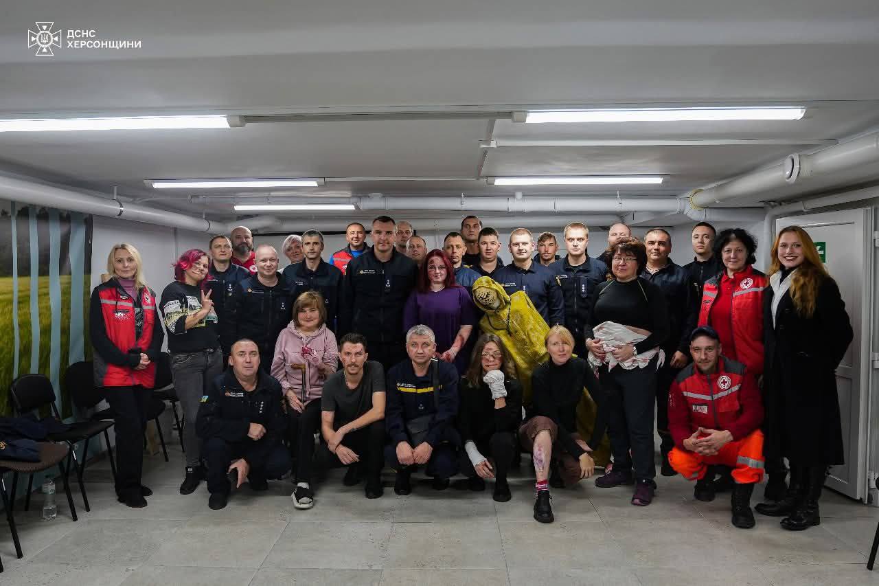 Групове фото близько 30 людей, серед яких є представники ДСНС Херсонщини, волонтери (деякі в червоних жилетах/одязі з емблемами Червоного Хреста) та інші цивільні особи. Вони стоять і сидять у приміщенні зі світлими стінами та лампами денного світла. У центрі уваги - жовтий захисний костюм, який, ймовірно, демонструється або використовується для навчання.