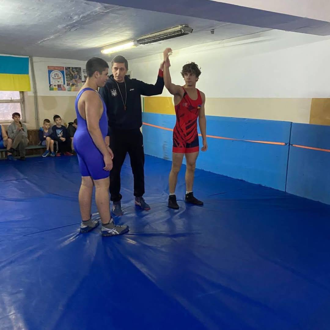 Троє людей на синьому борцівському килимі в спортивному залі. Суддя в центрі, одягнений у темне спортивне вбрання, піднімає руку борцю у червоно-синьому трико праворуч, оголошуючи його переможцем над борцем у синьому трико, що стоїть ліворуч. На задньому плані видно прапор України та сидять глядачі, переважно діти.