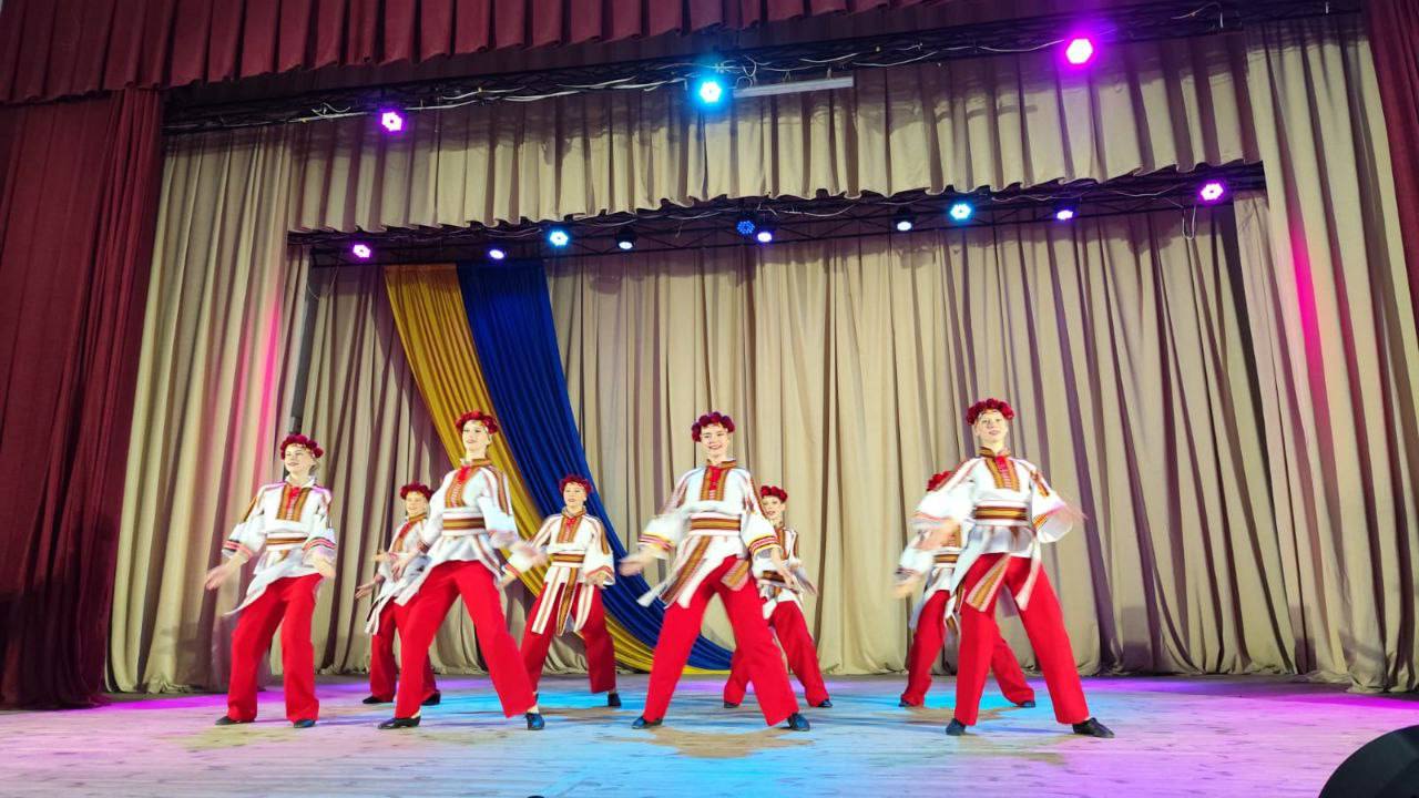 Група з семи молодих людей (жінок або дівчат) виконує танець на сцені. Вони одягнені в стилізовані українські народні костюми: вишиті білі сорочки з червоними елементами, червоні шаровари або штани та червоні вінки на головах. Сцена має важкі світло-коричневі/бежеві штори на задньому плані, прикрашені жовто-синіми (українськими) смугами тканини. Сцена яскраво освітлена синіми та фіолетовими прожекторами.