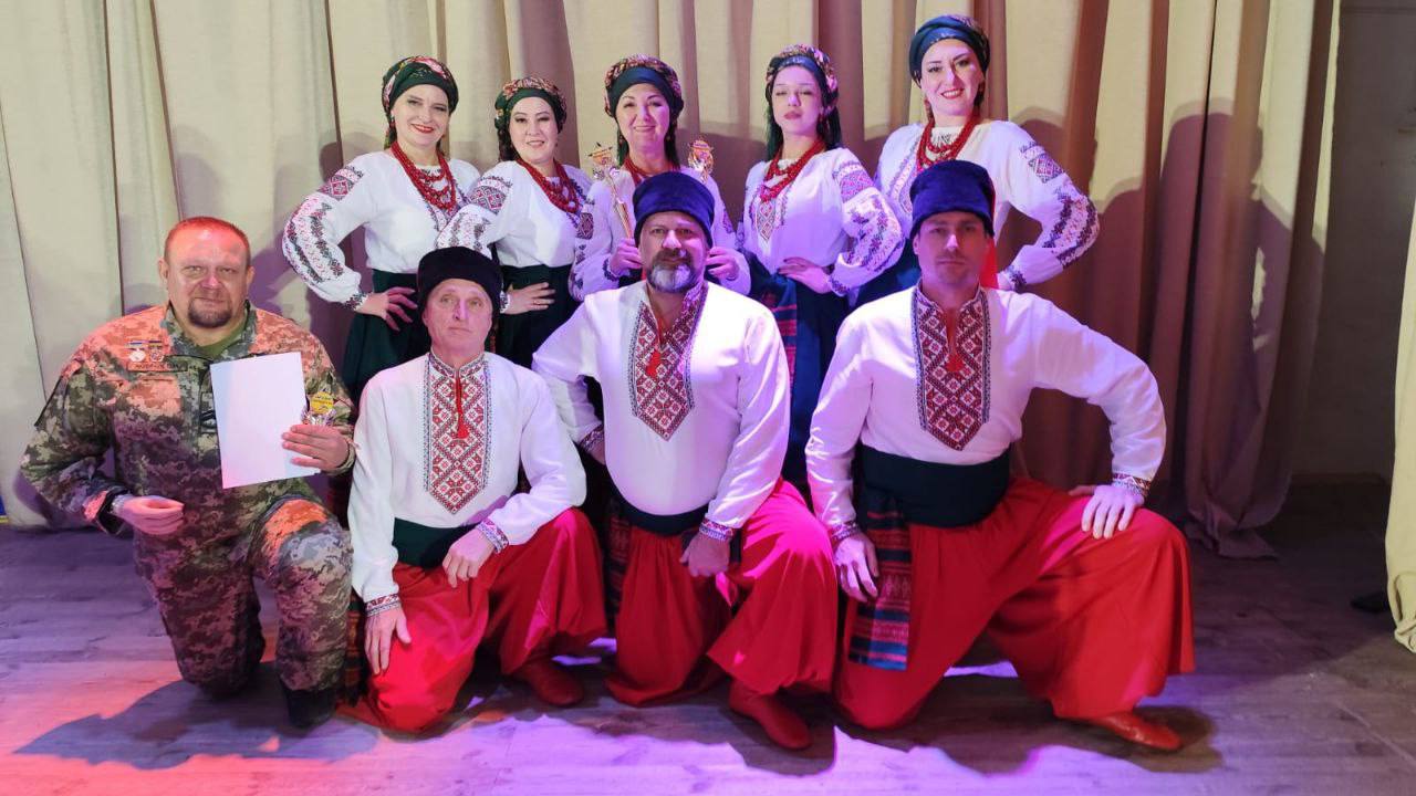 Групове фото восьми танцюристів у традиційному українському народному одязі та чоловіка у військовій формі на сцені перед світлими шторами. П'ять жінок стоять позаду, одягнені у вишиванки, з намистом та в хустках. Три чоловіки стоять або присідають спереду, одягнені у вишиванки та широкі червоні шаровари. Чоловік ліворуч одягнений у камуфляжну форму, тримає в руці білий аркуш паперу, ймовірно, це фото з концерту чи виступу.