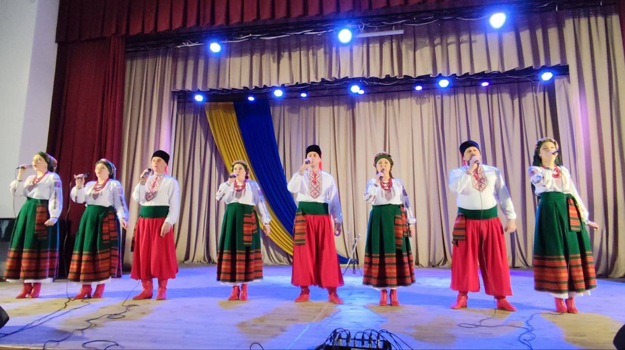 Група з семи виконавців (чотири жінки та три чоловіки) співає на сцені в традиційних українських народних костюмах. Жінки одягнені у вишиті блузки та довгі зелені спідниці з червоними елементами, чоловіки — у вишиті сорочки та червоні шаровари, а також у чорні головні убори. Усі тримають мікрофони. Сцена прикрашена світлими шторами ззаду та жовто-синіми (українськими) смугами тканини. Сцена освітлена синіми прожекторами.