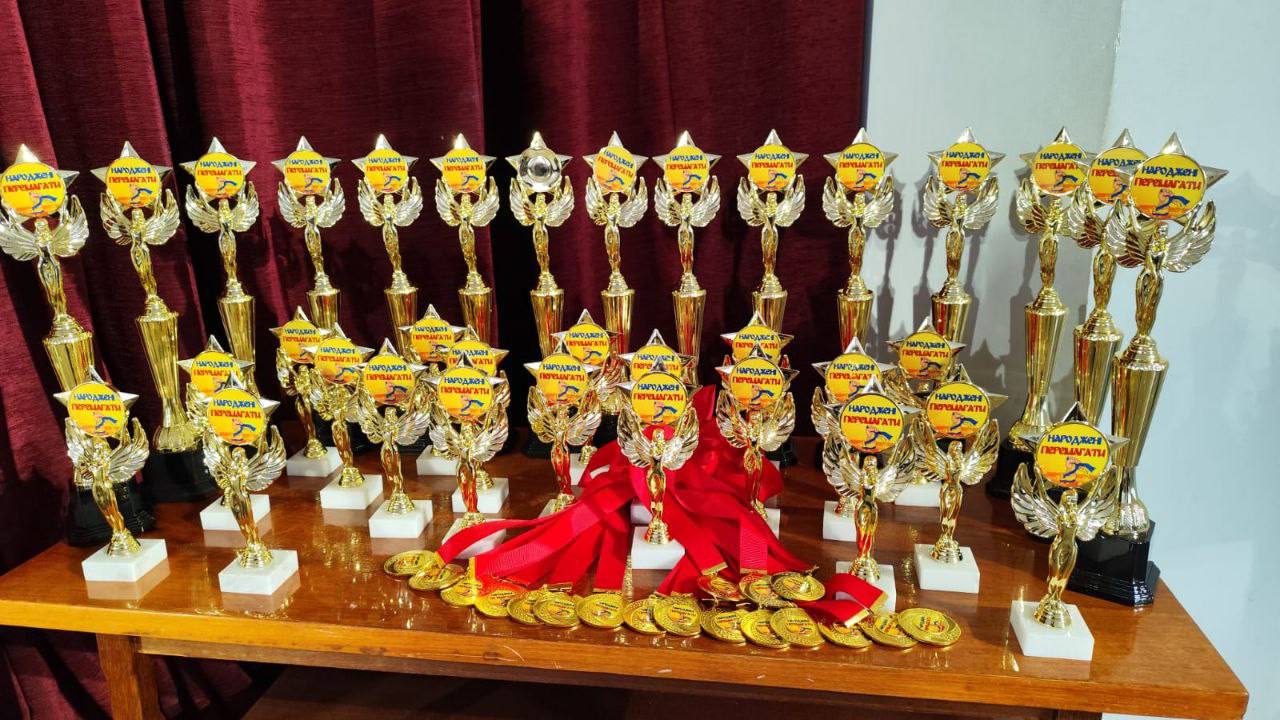 Горизонтальне фото відображає велику колекцію золотих трофеїв (кубків) і медалей, виставлених на дерев'яному столі, ймовірно, для церемонії нагородження. Кубки мають золотистий колір, стоять на білих або чорних основах і прикрашені елементами у вигляді зірок, крил та круглими емблемами з написами, які включають слово