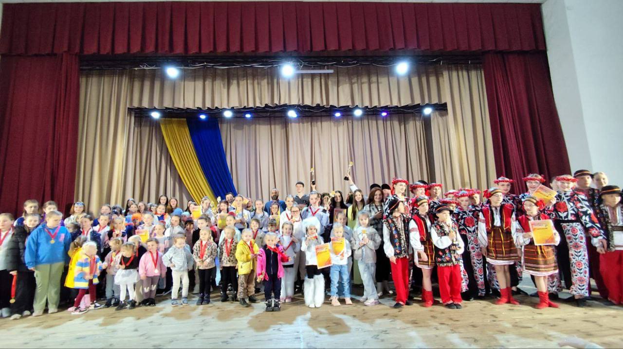 Велика групова фотографія на сцені, що зображує багато дітей, підлітків та дорослих, які брали участь у конкурсі чи фестивалі. Близько сотні людей стоять рядами, частина з них у барвистих українських народних костюмах, інші — у повсякденному одязі, з медалями, кубками та дипломами. На задньому плані сцени видно червоні та бежеві/світло-коричневі штори, а також синьо-жовтий (український) прапор або смуги тканини.