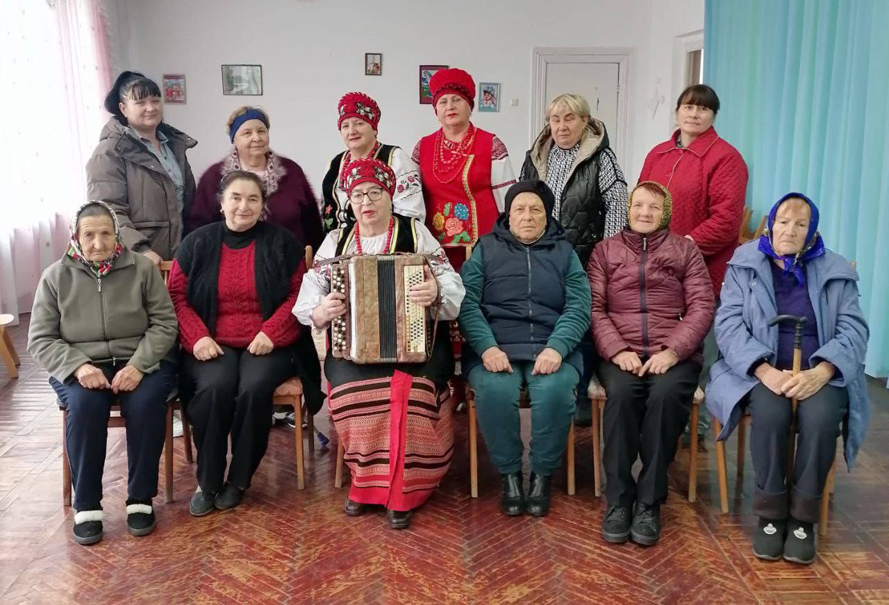 Фотографія групи з одинадцяти жінок у два ряди, знята у світлому приміщенні з дерев'яною підлогою. У першому ряду сидять п'ять літніх жінок, у центрі — виконавиця в яскравому українському народному костюмі з червоним головним убором, яка тримає гармошку (баян). Жінки у другому ряду стоять, частина з них також одягнена у стилізовані народні костюми.