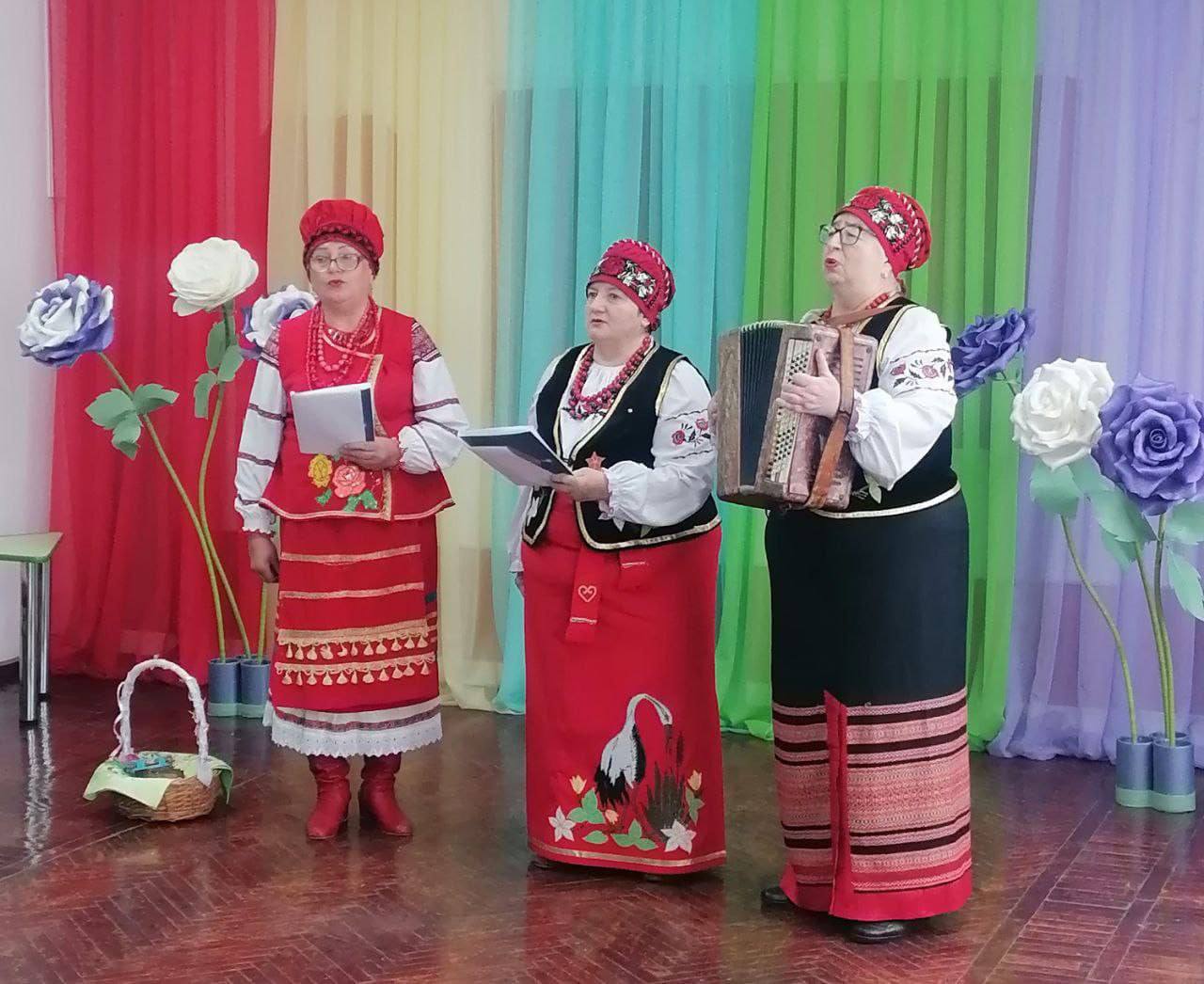 Три жінки, одягнені у вишиті білі сорочки, червоні плахти, корсетки та червоні головні убори, виступають на сцені. Жінки ліворуч і в центрі тримають нотні зошити, а жінка праворуч — грає на гармошці (баяні). Сцена оформлена яскравими вертикальними шторами (червоний, бірюзовий, зелений, рожевий) та великими декоративними трояндами синього, фіолетового та білого кольорів. На підлозі ліворуч стоїть плетений кошик.