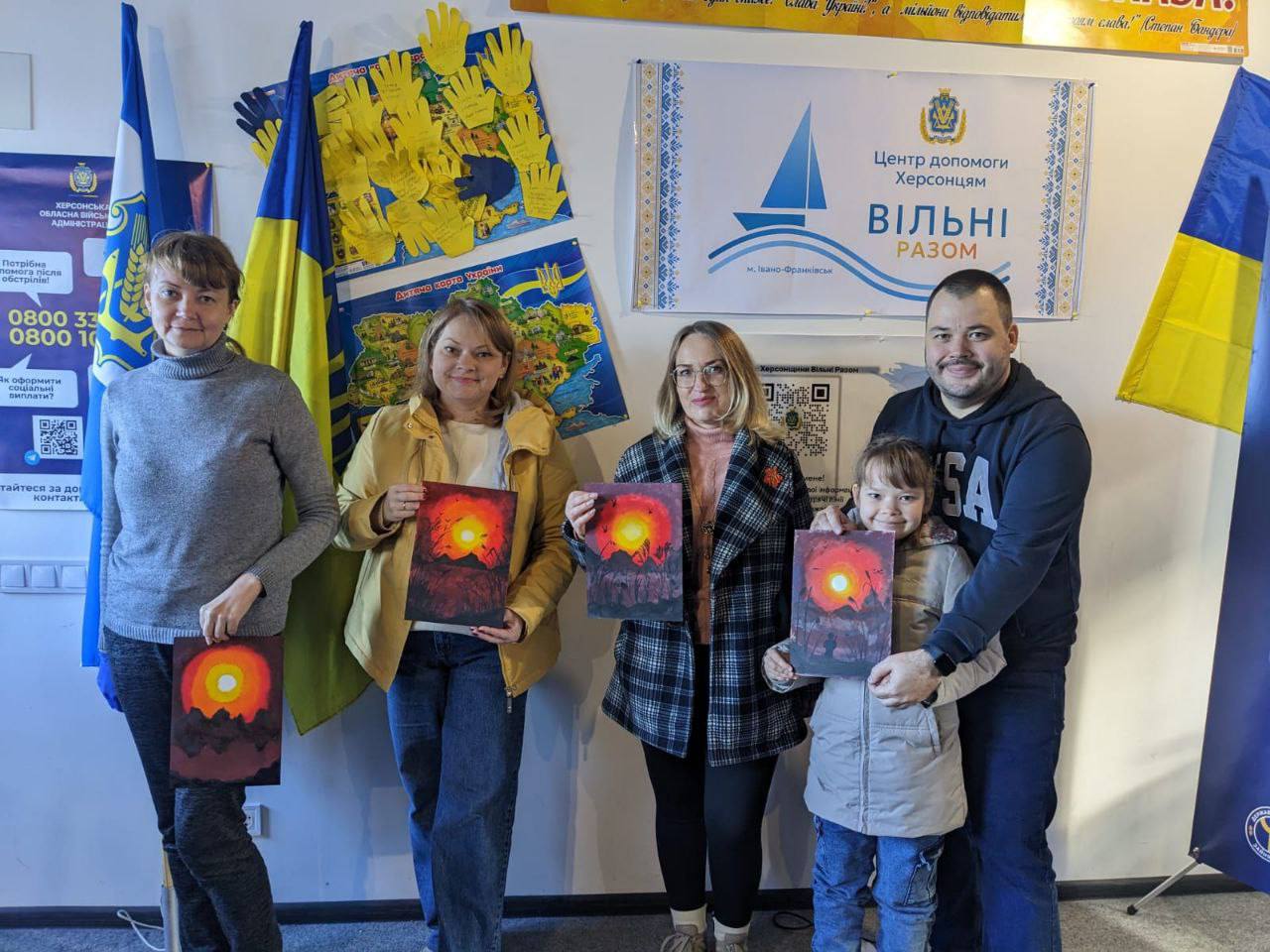 Групова фотографія п'яти осіб, які демонструють свої картини, виконані акрилом або олійними фарбами, із зображенням палаючого заходу сонця чи виверження вулкана. На фоні видно національні прапори України, банер «Центр допомоги херсонцям