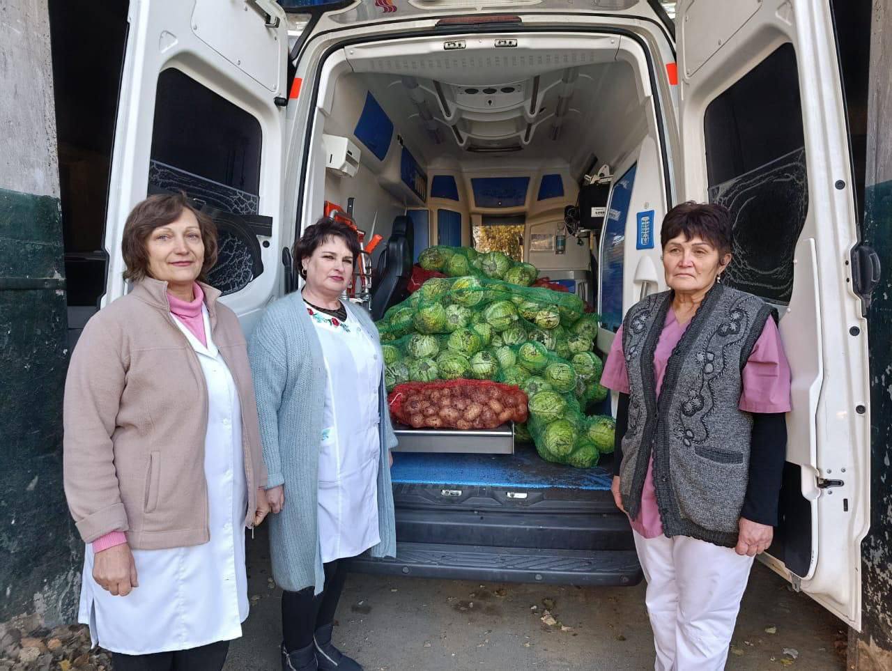 Три жінки у медичних халатах та одязі, що нагадує уніформу, стоять біля відчиненого заднього відсіку білого фургона. Всередині фургона лежить велика купа свіжої капусти в сітках, а також сітка з картоплею або цибулею.