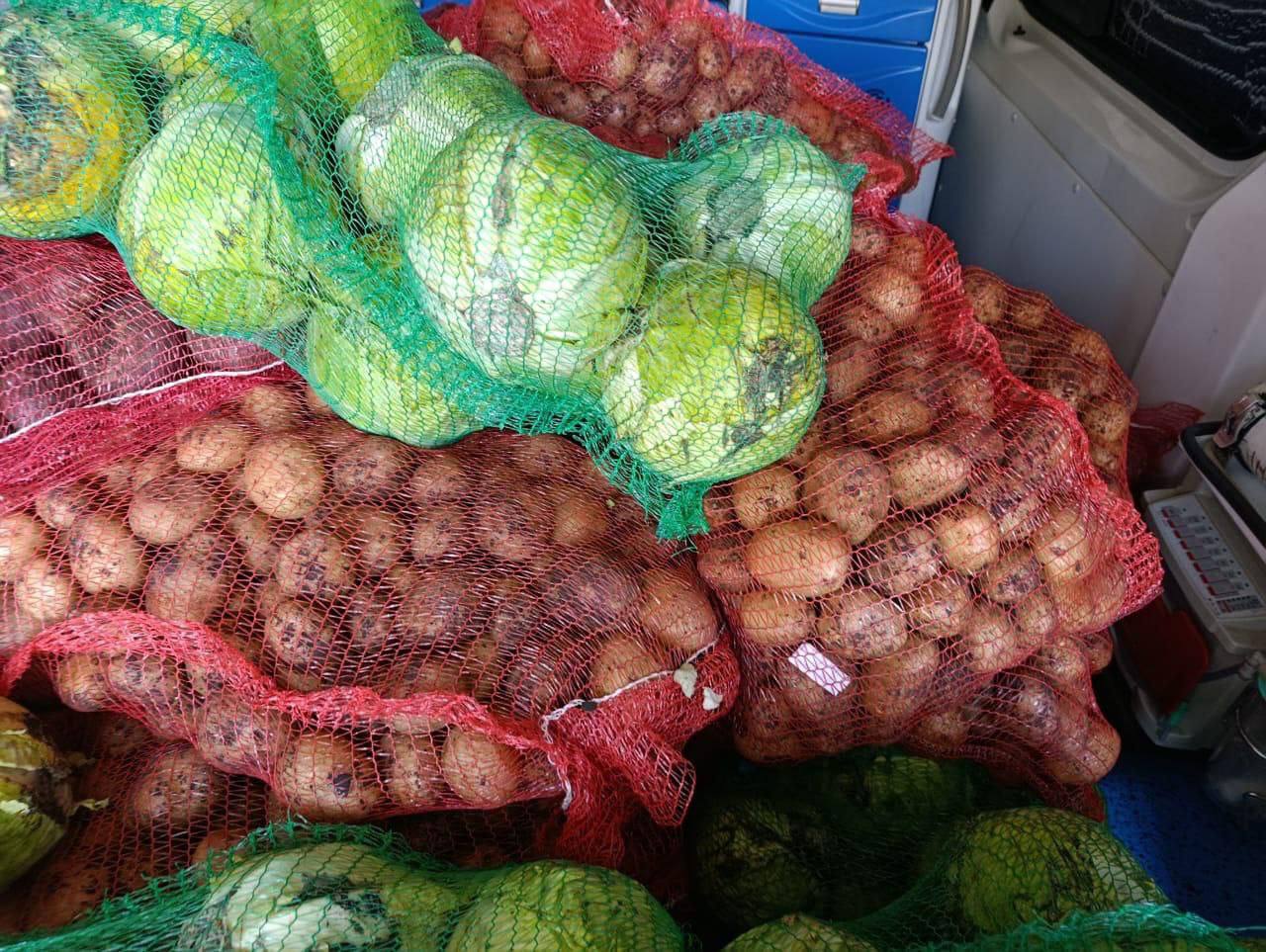 Крупний план заднього відсіку фургона або кузова, заповненого великими сітками з овочами. У кадрі домінує зелена капуста у зелених сітках та картопля у червоних сітках. Овочі викладені у велику купу.