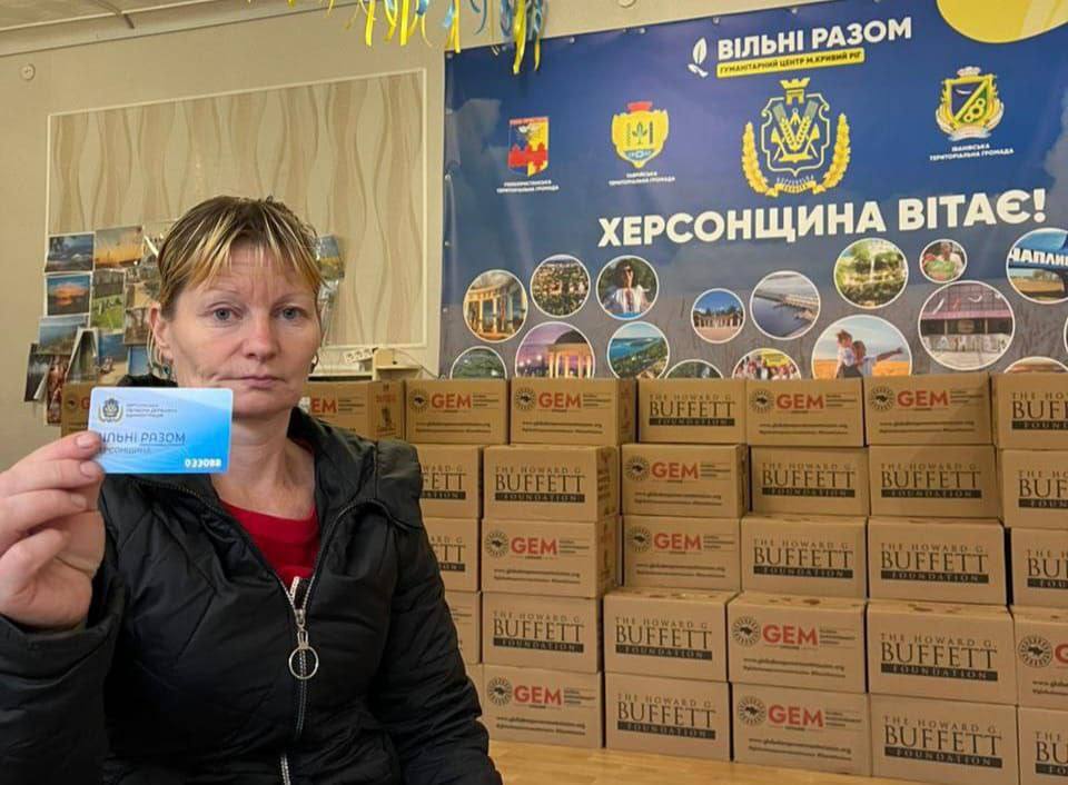 Жінка зі світлим волоссям у чорній куртці тримає в руці синю картку з написом 'ВІЛЬНІ РАЗОМ' та логотипом Херсонської області. Вона стоїть на тлі великого штабеля картонних коробок із логотипами The Howard G. Buffett Foundation та GEM, а позаду — синій банер 'ВІЛЬНІ РАЗОМ. ХЕРСОНЩИНА ВІТАЄ!' з гербами та фотографіями місцевості.