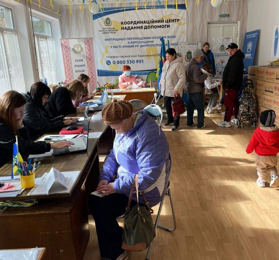Сцена у приміщенні Координаційного центру надання допомоги: волонтери за столами реєструють внутрішньо переміщених осіб з Херсонщини. На задньому плані розташований великий банер 