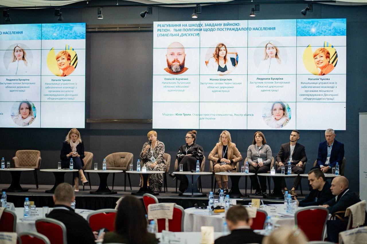 Панельна дискусія "Реагування на шкоду, завдану війною: регіон — цілі політики щодо постраждалого населення" (заголовок на екрані). На сцені сидять шестеро жінок і двоє чоловіків (усі в цивільному одязі), які беруть участь в обговоренні. Одна з учасниць ліворуч тримає мікрофон. За ними розташований великий екран, на якому відображені імена та посади учасників.