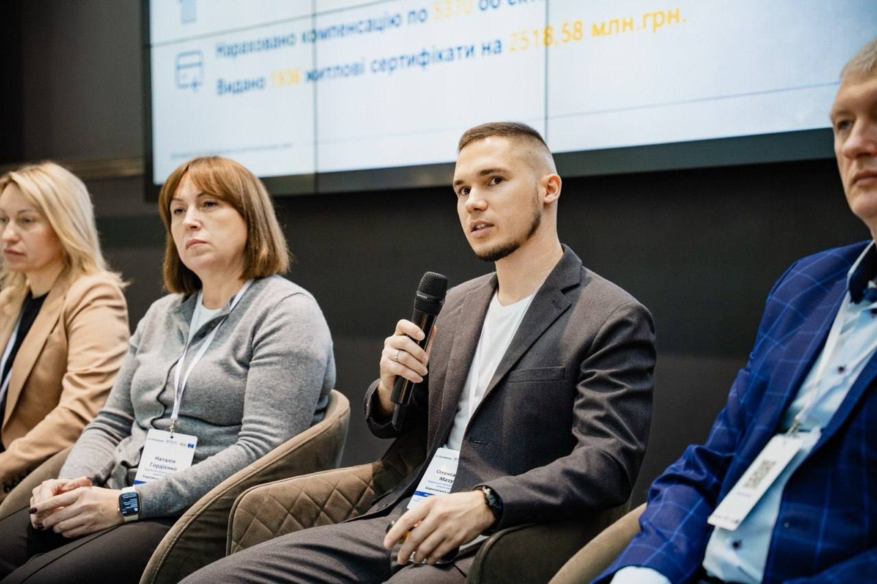 Груповий знімок спікерів на панельній дискусії. На передньому плані молодий чоловік у сірому костюмі говорить у мікрофон. Ліворуч від нього сидять дві жінки, на бейджі однієї з них видно ім'я Наталія Гордієнко. На задньому плані – розмитий екран із даними про компенсацію та видані житлові сертифікати.