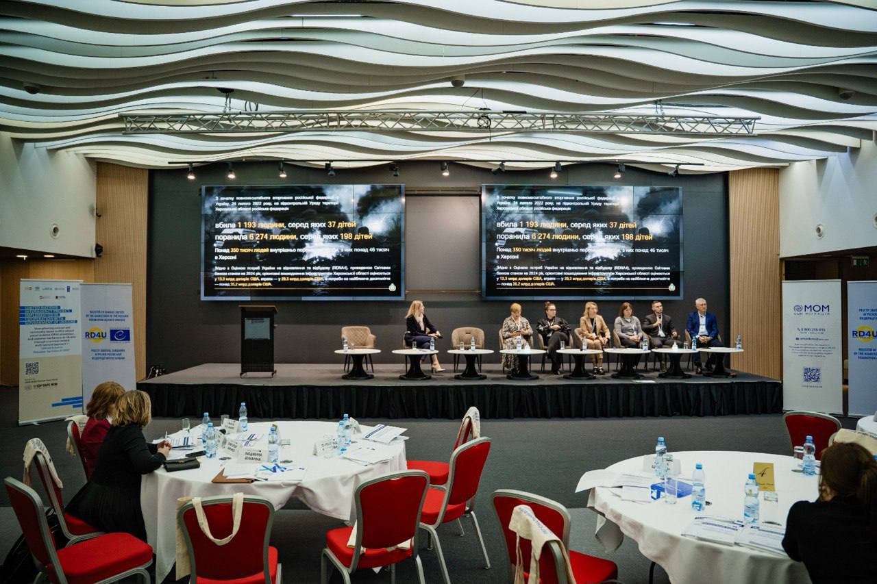 Загальний план конференц-залу з панельною дискусією на сцені. Сім спікерів сидять за столиками на підвищенні. Над ними два великі екрани з ідентичною статистикою втрат серед населення, включаючи дані про загиблих (1193) та поранених (6274), а також внутрішньо переміщених осіб. На передньому плані видно круглі столи з червоними стільцями та присутніх учасників.