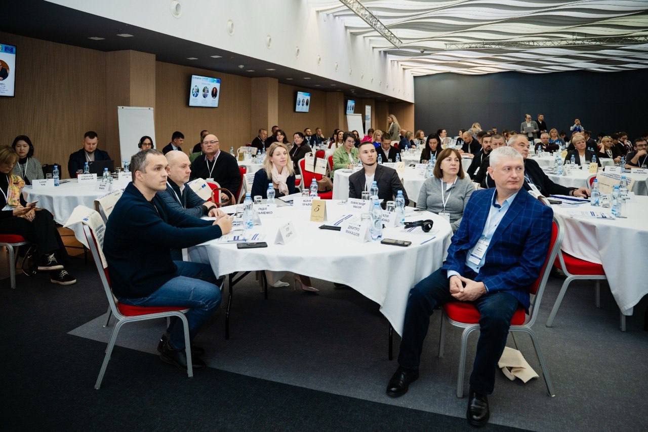 Фотографія аудиторії конференції. Учасники сидять за круглими столами на червоних стільцях, слухаючи виступ. Зал заповнений людьми, на столах розташовані пляшки з водою та матеріали. Інтер'єр залу має сучасний дизайн із хвилястою стелею.