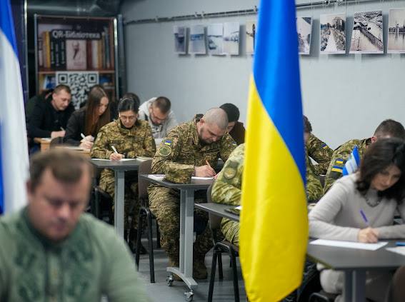 Група людей сидить за партами у класі чи аудиторії, зосередившись на написанні тексту чи заповненні документів. Деякі учасники одягнені у військову камуфляжну форму ЗСУ, інші — у цивільному одязі. На передньому плані праворуч розміщено великий прапор України (синьо-жовтий), а ліворуч — частина іншого прапора. На задньому плані видно стелаж із книгами, плакат із написом "QR-Бібліотека" та низку чорно-білих фотографій на стіні.