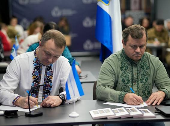 Двоє чоловіків сидять за столом і зосереджено пишуть. Чоловік ліворуч одягнений у білу вишиванку з яскравою вишивкою, чоловік праворуч — у світло-зелену вишиванку. На столі перед ними лежать папери, ручки, мініатюрні синьо-білі прапорці та стопка марок або невеликих карток. На задньому плані видно синьо-білий прапор та розмиті фігури інших учасників.
