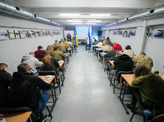 Широкий кадр із навчального приміщення (можливо, бомбосховища або підвалу) з низькою стелею, де відбувається заняття. Учасники, включно з військовослужбовцями у камуфляжі та людьми у цивільному, сидять за партами, розташованими у два довгі ряди. У кінці аудиторії видно екран, прапори України (синьо-жовтий) та інший (синьо-білий), а також викладачів, які стоять перед групою. На стінах з обох боків висять ряди чорно-білих і кольорових фотографій.