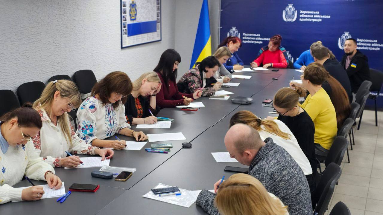 Група людей, переважно жінок, сидять за довгим столом у приміщенні та пишуть, зосередившись на паперах перед собою. Кілька жінок одягнені у вишиванки. На столі видно ручки, мобільні телефони та папери. На задньому плані розташований прапор України (синьо-жовтий), а також синій банер (пресвол) із логотипами та написом "Херсонська обласна військова адміністрація".