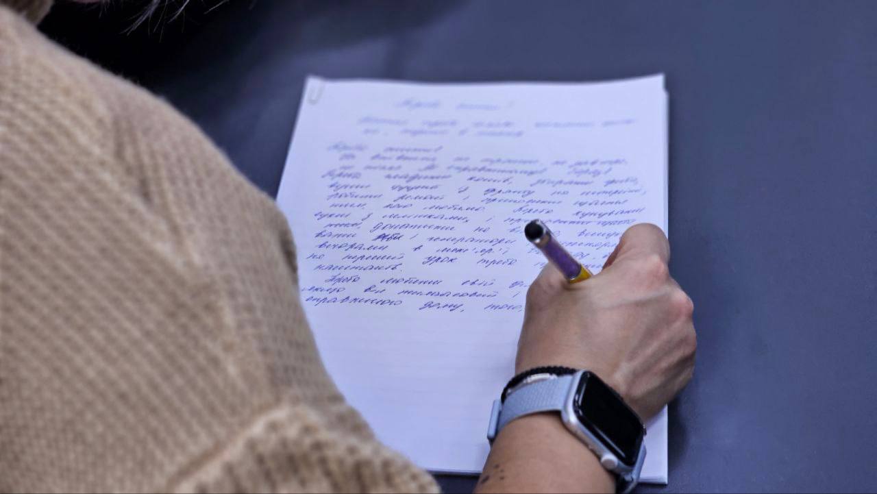 Крупний план руки людини в світлому в'язаному светрі, яка пише ручкою з фіолетовим корпусом на аркуші паперу, заповненому рукописним текстом українською мовою. На зап'ясті видно наручний годинник або фітнес-трекер зі світло-сірим ремінцем.