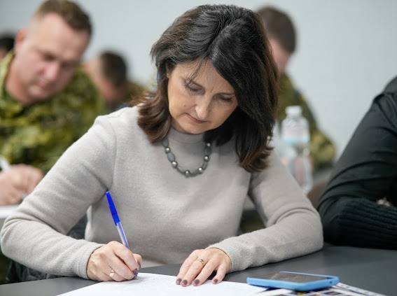 Жінка середнього віку з темним волоссям у світло-сірому светрі та сірому намисті сидить за столом і зосереджено пише синьою ручкою на аркуші паперу. Поруч на столі лежить мобільний телефон у синьому чохлі. На задньому плані видно розмиті фігури інших учасників, включно з чоловіками у камуфляжній формі.