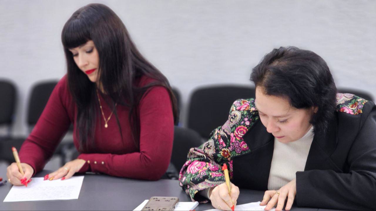 Дві жінки сидять за столом, зосереджено пишучи ручками на аркушах паперу. Жінка ліворуч, з довгим темним волоссям і яскраво-червоною помадою, одягнена у бордовий светр. Жінка праворуч одягнена у чорний піджак із яскравими квітковими вставками на плечах поверх світлого гольфа. На столі видно мобільний телефон.